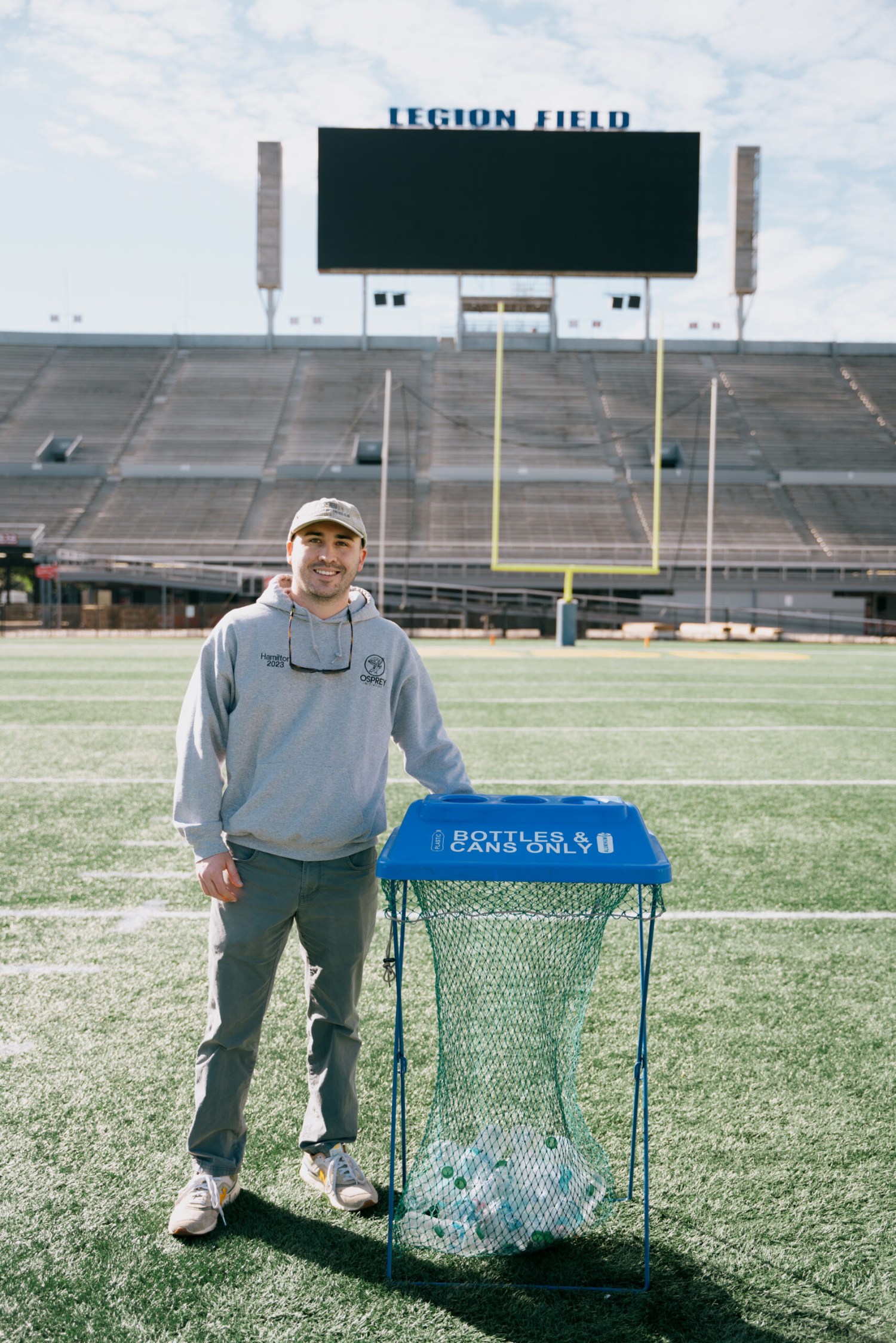 Man standing beside a bottles and cans recycling bin on the field at Legion Field stadium