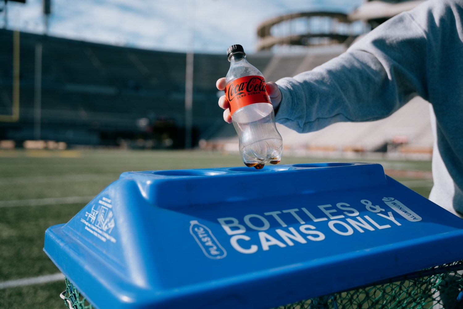 Person placing an empty Coca-Cola bottle into a stadium recycling bin labeled "Bottles & Cans Only